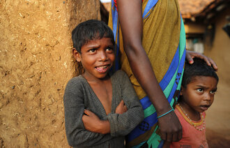 Bildet er fra India og viser to barn som står sammen med en voksen person og ser på helsearbeidere som overtaler eldre til å vaksinere seg mot korona