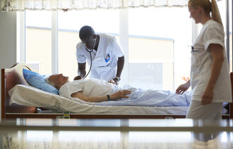 Bildet viser en sykepleier som lytter med stetoskop på en eldre mann i sengen. Ved siden av står en annen sykepleier og observerer.