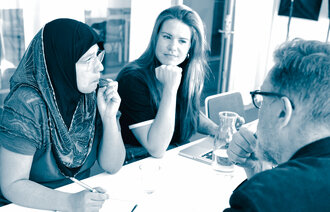 The photo shows a woman wearing a hijab, a woman wearing Western clothes and a man in a suit, sitting around a table. They are attending a meeting.