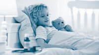 The photo shows an ill, little boy lying in his bed. In his handen he is holding a teddy bear. There are medicine bottles on the bedside table.