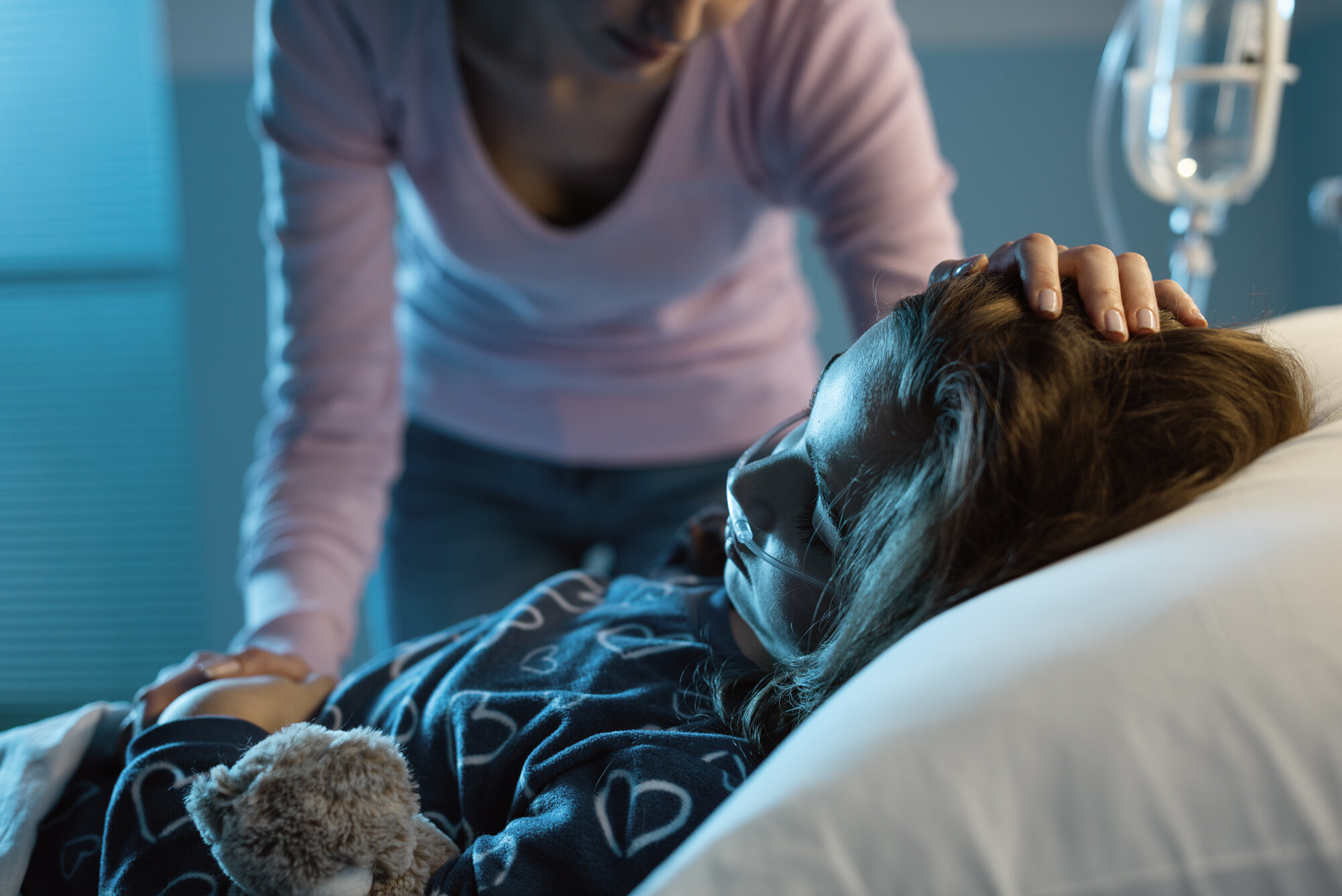 Bildet viser ei syk jente i senga på sykehuset. Moren står over og holder henne på pannen.