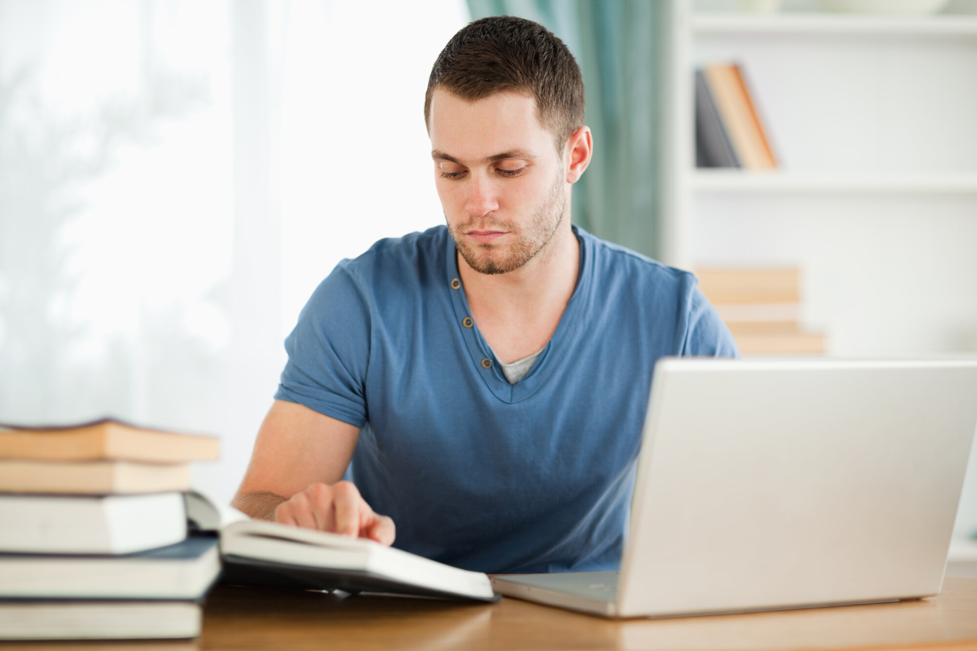 Bildet viser en mannlig student som leser i bøker og skriver på en laptop