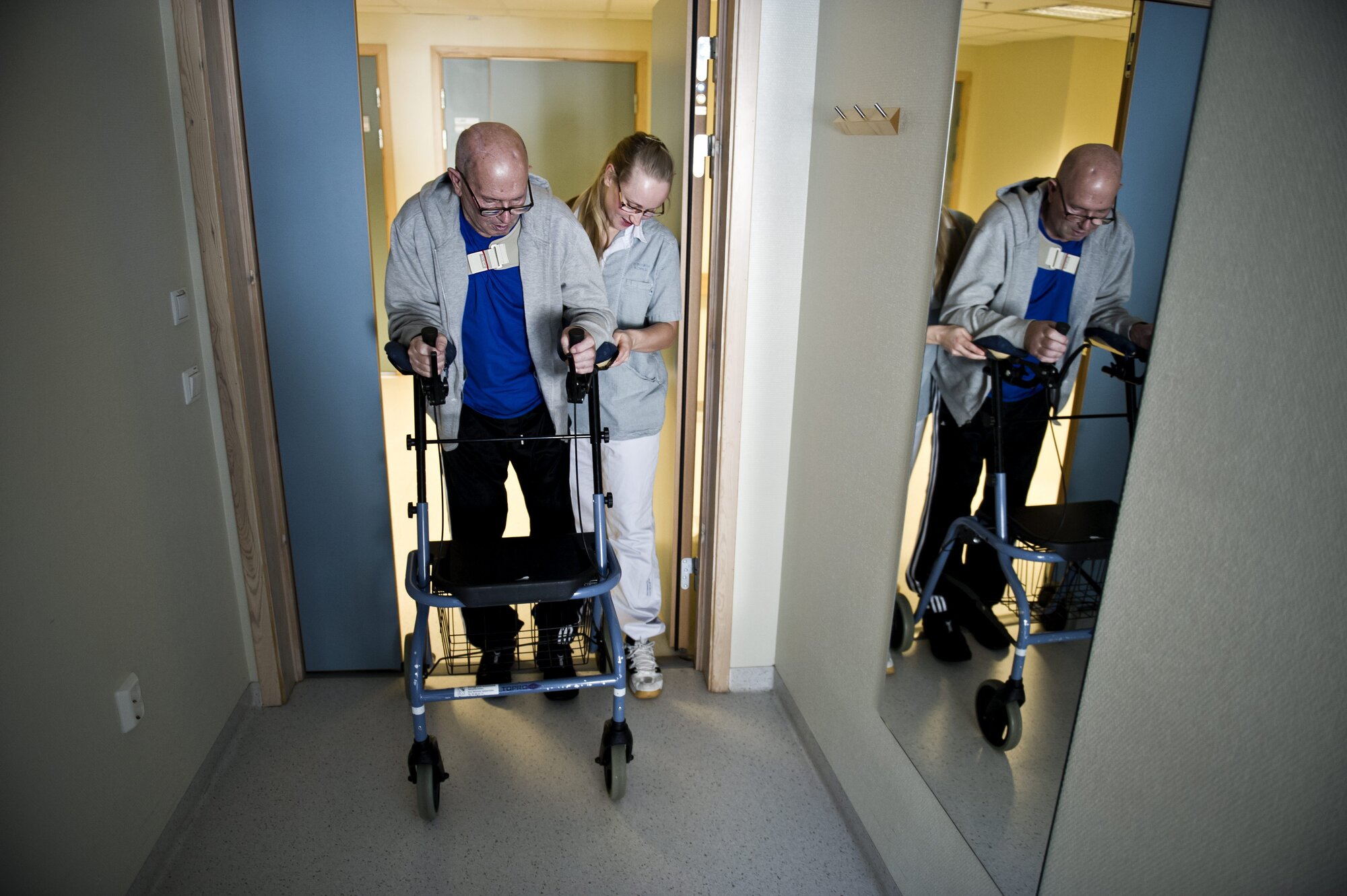 Bildet viser en mann med gåstol, som støttes av en sykepleier