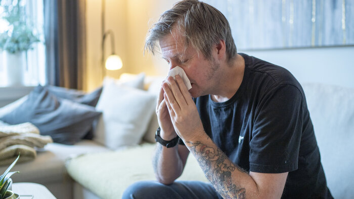 Bildet viser en mann som sitter i en sofa og snyter seg.