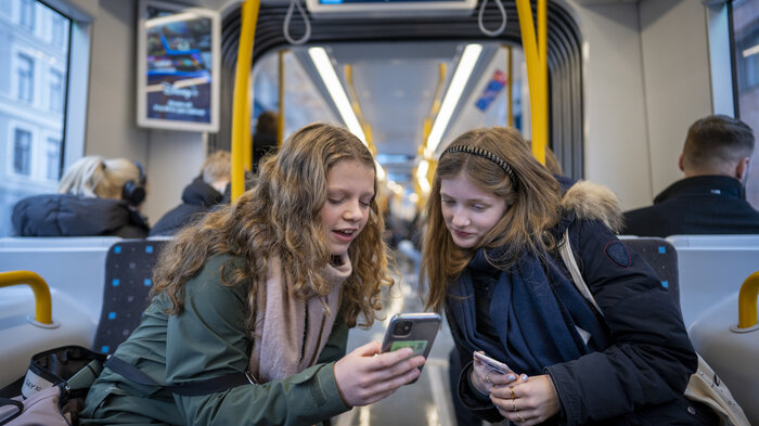 Bildet viser to unge jenter som ser på mobilene sine på trikken.