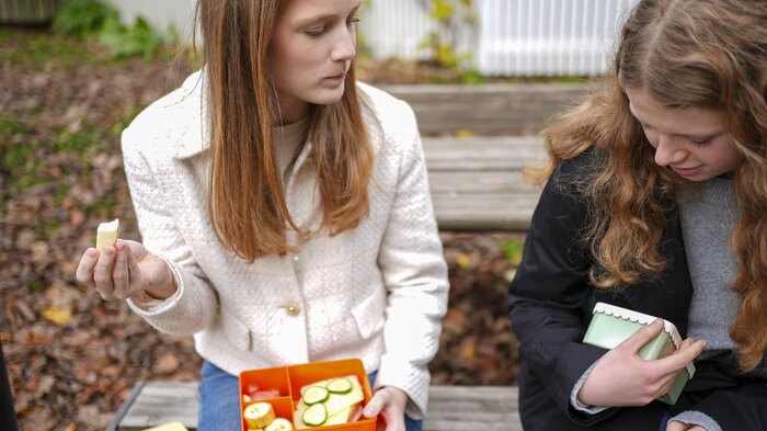 Bildet viser to jenter som spiser lunsj på en benk.