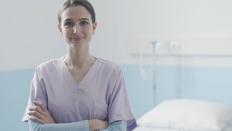 Bildet viser en ung sykepleier som står med armene i kors foran en sykehusseng