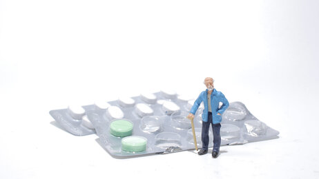 Bildet viser en figur av en eldre mann som står ved siden av et brett med medisiner