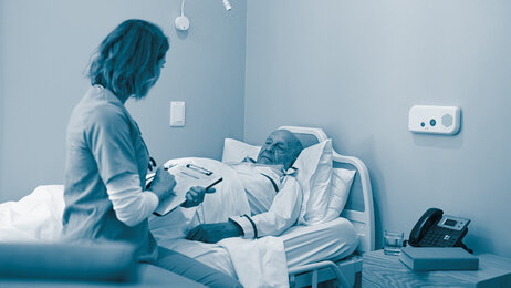 The photo shows a nurse sitting by the bed of an elderly man. She is holding a form in her hand.