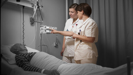 Bildet viser to sykepleiere som står ved en pasientseng