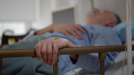 The photo shows an older man lying in a hospital bed. His left hand is resting on the bed rail. In his hand is a venous access port.