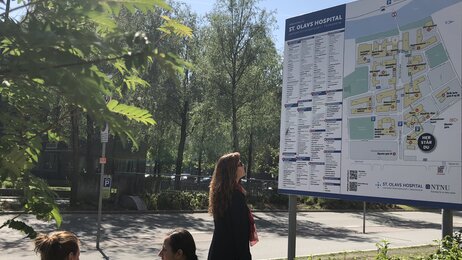 The picture shows a woman standing in front of a large sign outside St. Olavs Hospital. The sign displays information about the hospital.