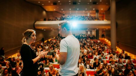 Bildet viser to studenter som står på scenen i et auditorium