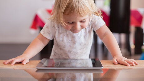 Bildet viser en smilende, liten jente som står og kikker ned på er nettbrett som ligger på bordet