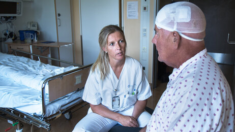 Bildet viser en sykepleier som sitter overfor en nyoperert parkinsonpasient. Han har bandasje rundt hodet