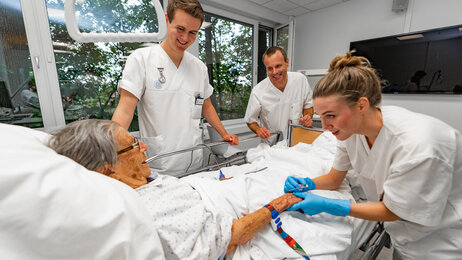 Bildet viser en eldre pasient som ligger i en sykehusseng. Rundt henne står to sykepleierstudenter og en veileder. Alle smiler, og den ene studenten holder pasientens hånd