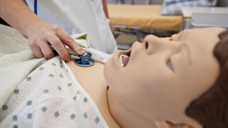 Bildet viser en hånd som holder et stetoskop på brystet til en simuleringsdukke