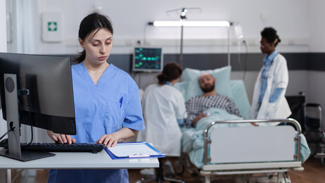 Bildet viser en sykepleier som står på et pasientrom foran en PC. Foran henne ligger et skjema. Bak henne ligger en pasient i senga med to sykepleiere ved sengekanten