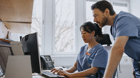 Sykepleiere foran PC Bildet viser en kvinnelig sykepleier som sitter foran en PC. En mannlig sykepleier står bak henne og kikker på skjermen