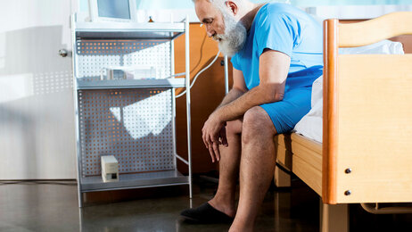Bildet viser en eldre mann med skjegg som sitter på kanten av en sykehusseng med hendene i fanget.
