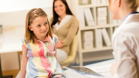 Bildet viser en liten jente som sitter på pulten til en helsesøster. Moren sitter i bakgrunnen.