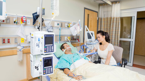 Bildet viser en mannlig pasient som ligger i sengen på sykehus. En kvinnelig pårørende sitter i en stol ved siden av.