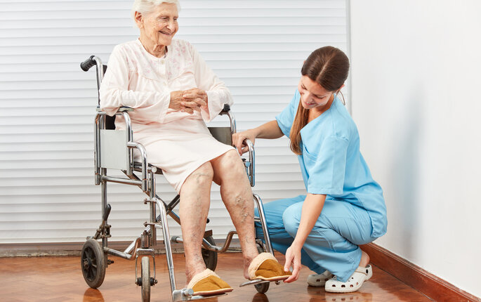 Bildet viser en sykepleier som sitter på golvet og hjelper en eldre dame i en rullestol.