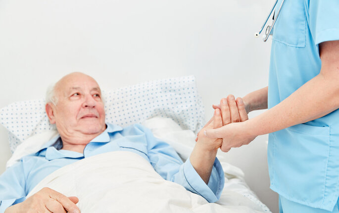 Bildet viser en eldre mann som ligger i senga. En sykepleier står ved siden av og holder ham i hånda