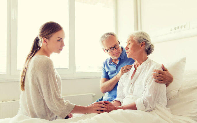 Bildet viser en eldre, syk dame i senga. Familien sitter på sengekanten, og en mann holder rundt henne.