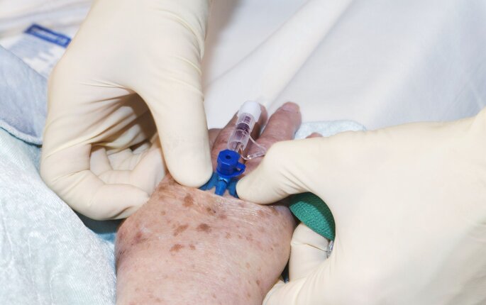 Bildet viser hendene til en sykepleier, som holder på å legge inn perifert venekateter på en hånd til en eldre person