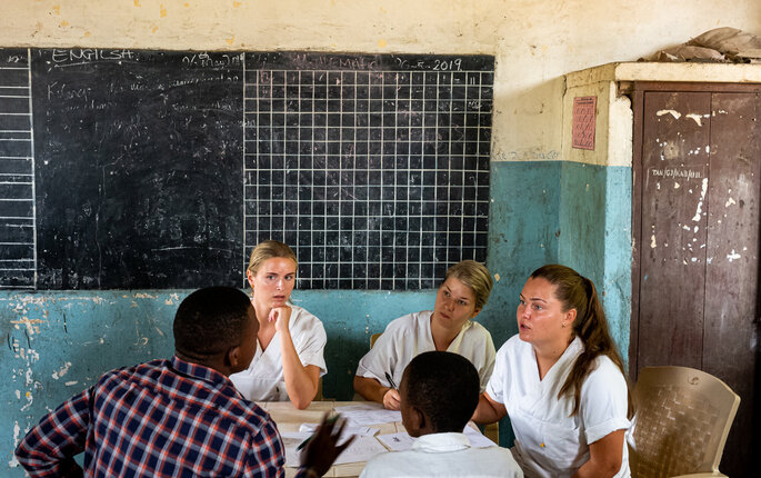 Bildet viser tre sykepleierstudenter fra Norge som er i Tanzania. De sitter og snakker med to afrikanske menn.