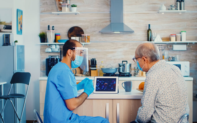 Bildet viser en mannlig sykepleier i blå uniform, munnbind og visir på hjemmebesøk hos en eldre mann, også han med munnbind. De sitter og ser på en ipad.