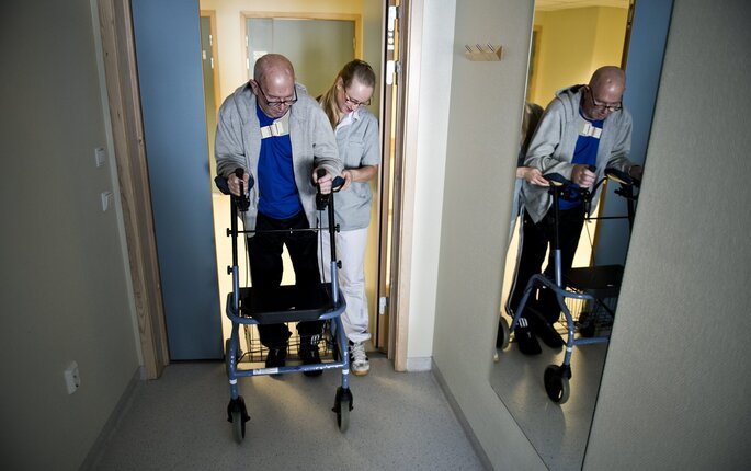 Bildet viser en mann med gåstol, som støttes av en sykepleier