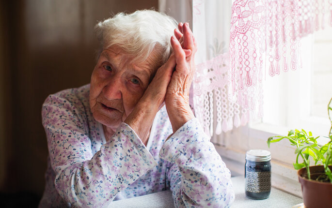 The photo shows an old, sad woman holding her head in her hands. On the table in front of her is a glass of pills.