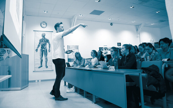 The photo shows a male lecturer in an auditorium full of female students