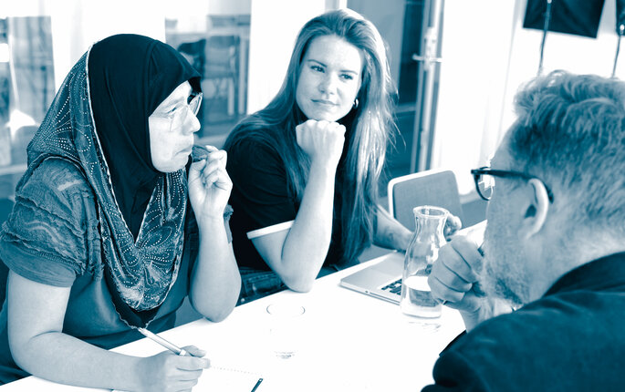 The photo shows a woman wearing a hijab, a woman wearing Western clothes and a man in a suit, sitting around a table. They are attending a meeting.