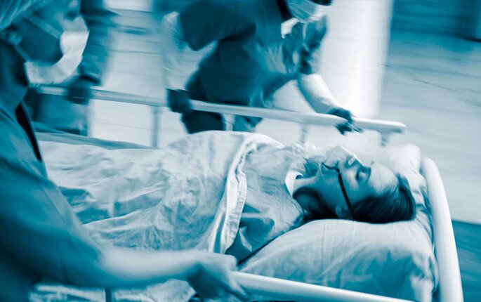 The photo shows a patient on a stretcher carried by running health professionals. The patient is wearing an oxygen mask.