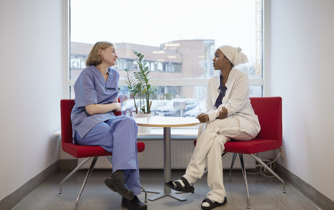 The photo shows two nurses sitting and talking together in front of a window