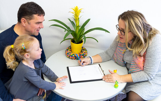 The photo shows a father with his daughter on his lap.  They are sitting at a table opposite a woman leaning towards them. She has a notebook in front of her.