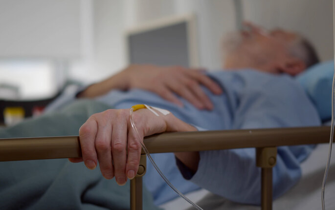The photo shows an older man lying in a hospital bed. His left hand is resting on the bed rail. In his hand is a venous access port.