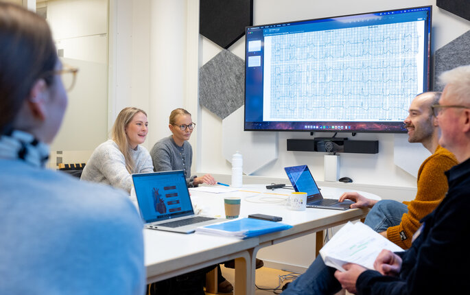The photo shows two students sitting around a table opposite two academic supervisors. A third student can be seen with her back towards the camera. At the screen on the wall graphs of cardiac arrhythmias can be seen. The people are participating in a reflection group.
