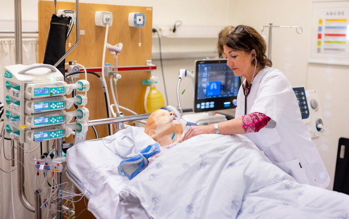 Bildet viser en simuleringsdukke på respirator. En sykepleier står lent over senga.