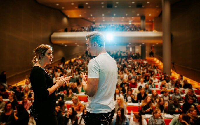 Bildet viser to studenter som står på scenen i et auditorium