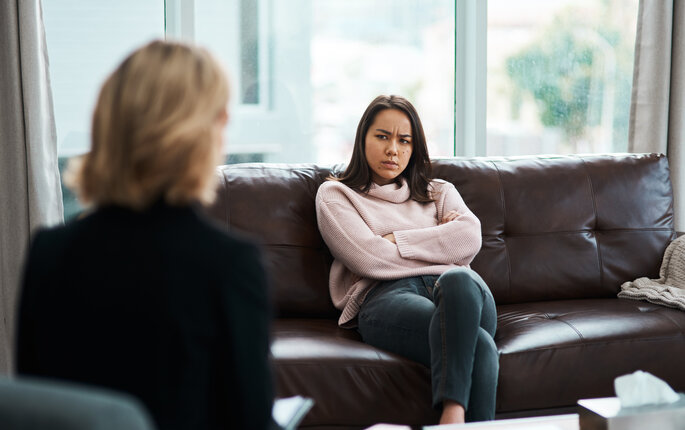 Bildet viser en person som sitter med ryggen til. Overfor henne sitter en ung jente som ser sint ut. Hun har armene i kors og et sint uttrykk