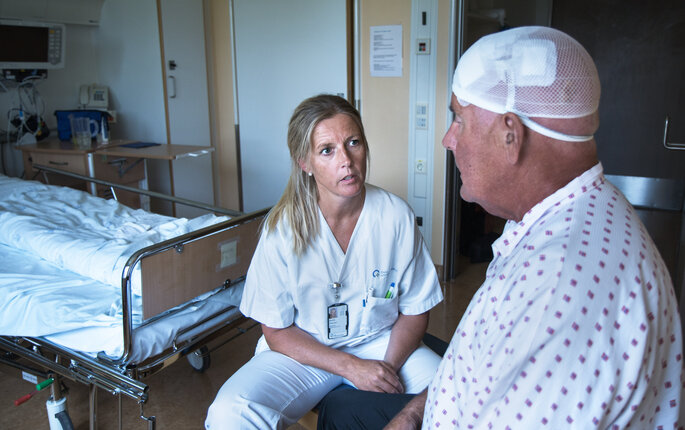 Bildet viser en sykepleier som sitter overfor en nyoperert parkinsonpasient. Han har bandasje rundt hodet