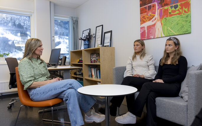 Bildet viser en helsesykepleier på kontoret som sitter i en stolgruppe overfor to ungdommer