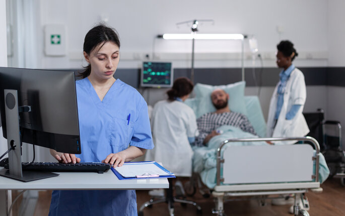 Bildet viser en sykepleier som står på et pasientrom foran en PC. Foran henne ligger et skjema. Bak henne ligger en pasient i senga med to sykepleiere ved sengekanten