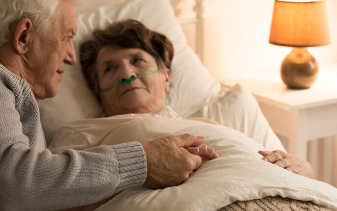 Bildet viser en syk eldre dame som ligger i senga med neseslange. En eldre mann holder henne i hånda og kikker på henne