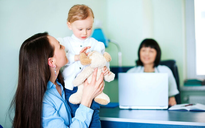 Bildet viser en mor som løfter sitt barn. De sitter foran pulten til helsesøsteren, som skimtes i bakgrunnen.