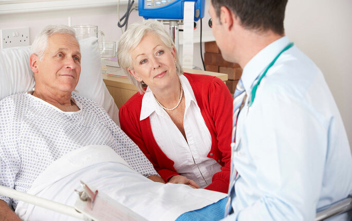 Bildet viser en eldre mann som ligger i senga på sykehuset. Kona sitter ved siden av, og legen er på visitt.